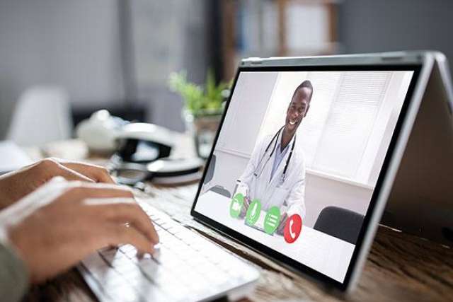 A doctor appears on a video call, smiling, while a person types on a laptop.
