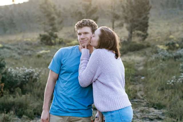 A young woman kisses her boyfriend on the cheek while they stand outdoors.