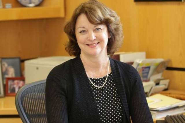 Person wearing a black cardigan over a black polka-dot dress with a pearl necklace, seated at an office desk with papers, folders, and framed photos in the background.