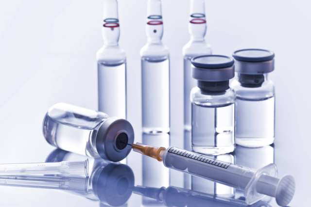 A syringe drawing clear liquid from a medical vial, with other vials and three ampoules visible in the background on a bright surface.