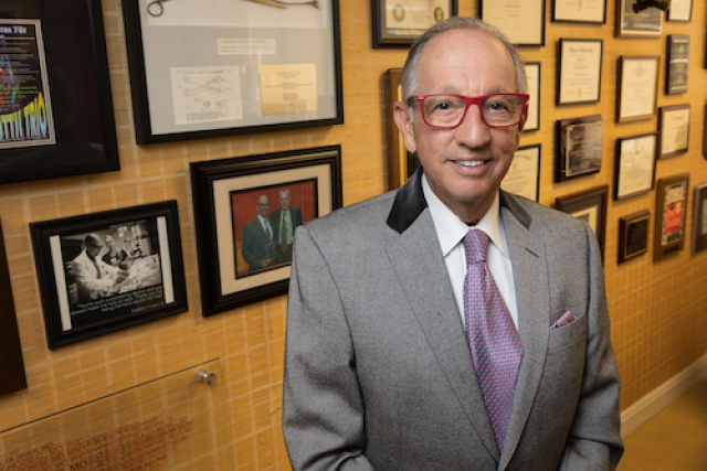 Ronald W. Busuttil, MD, PhD standing in front of a yellow wall with many picture frames, smiling and looking at the camera