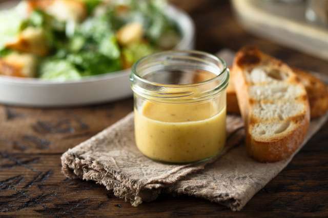 A jar of mustard dressing beside toasted bread on a wooden surface, with a salad in the background.