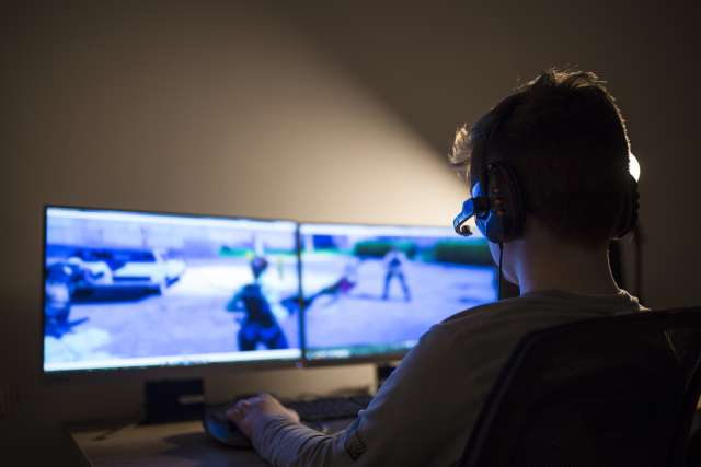 A boy plays a video game using two computer screens and a headset.