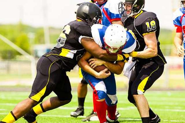 Football players in black and yellow uniforms tackling a player in blue and white on a green field.
