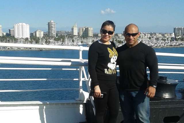 Two people standing on a boat deck with a city skyline and marina full of sailboats in the background, under a clear blue sky.