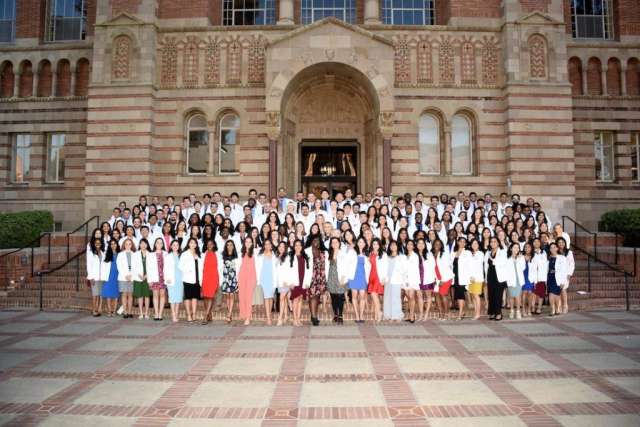 A large group of medical students stand in rows on the steps of a brick building, many wearing white coats.