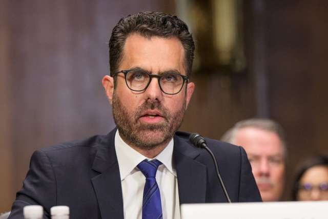 Man in suit and glasses speaking at a formal hearing behind a microphone and nameplate