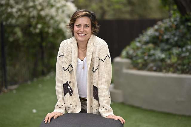 Melanie Gretsch wearing a cream cardigan with patterns stands outdoors among greenery.