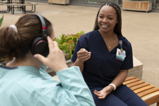  Shoni Taylor, wearing dark blue scrubs, smiles while talking to another woman on a bench outdoors. The other woman has headphones on.