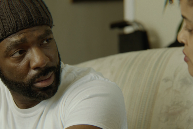 A bearded African American man in a dark knit cap and white t-shirt looks intensely at a person, whose face is partially visible on the right.