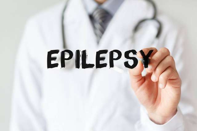 A doctor in a white coat and stethoscope writes the word "EPILEPSY" in bold black letters on a transparent surface with a white marker.