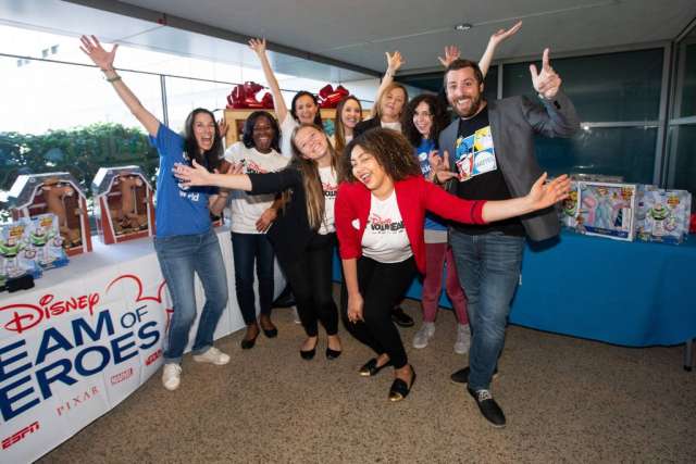 A group of 10 smiling people, some wearing Disney-themed clothing, pose excitedly with arms raised around a "Disney Team of Heroes" banner.