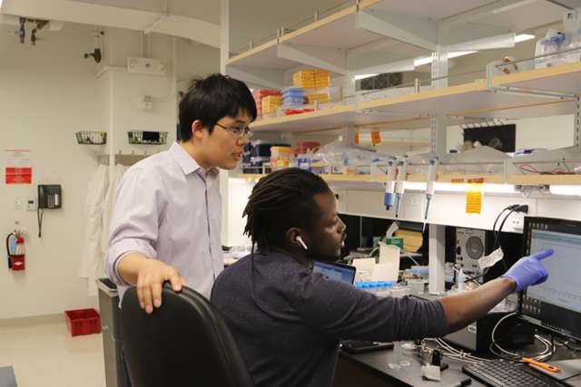 Two researchers at lab workstation, one operating equipment while the other observes.