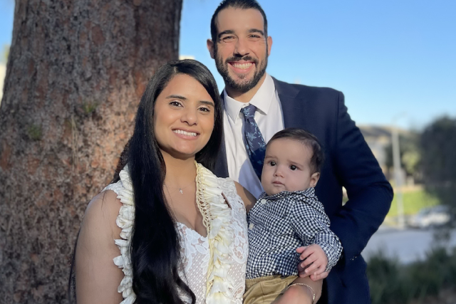 Dr. Cindy Hudson, UCLA Health OB-GYN, smiling outdoors with her partner and baby, dressed in formal attire on a sunny day.