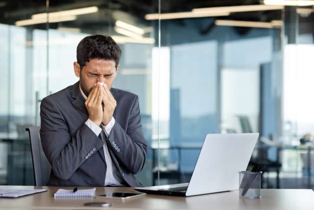 A man with allergies sneezes while at his work desk, covering his nose with a tissue.