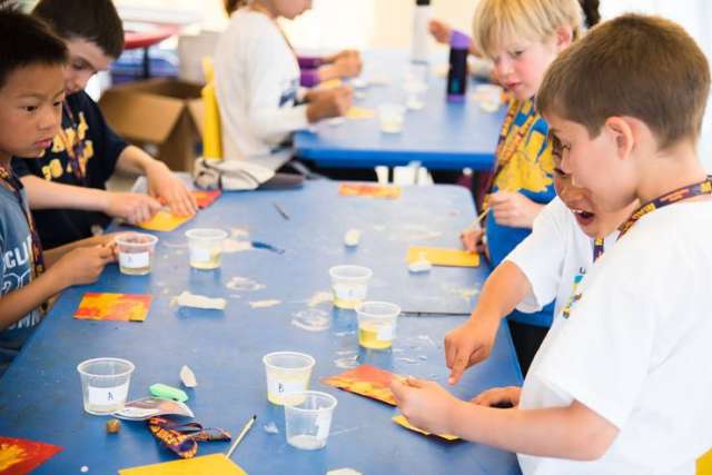 Children sit at a table doing crafts with cups, tools, and colorful paper during an indoor activity.