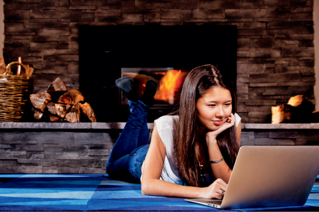 Teen girl lies on a rug in front of a fireplace, smiling while using a laptop.
