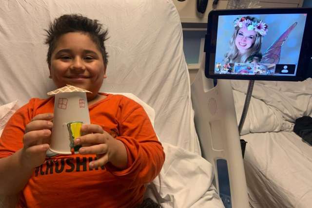 Child in a hospital bed holds a cup, with a digital screen displaying a smiling girl in a flower crown.