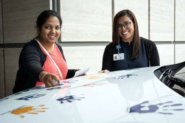  Two women smile while leaning over a white car hood adorned with colorful handprints. One points at the handprints.