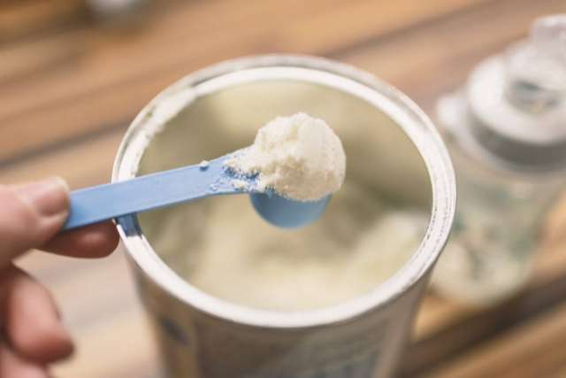Scoop of infant formula held above an open container with a baby bottle in the background.