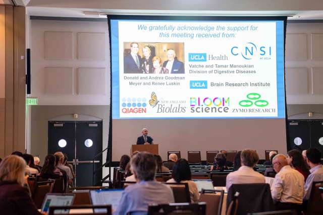 Conference presenter at podium with projected sponsor slide and seated audience.
