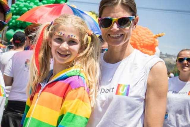 Two people at an outdoor event wearing rainbow-themed clothing, with colorful balloons and umbrellas in the background.