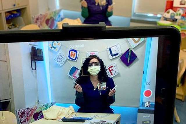 A woman in scrubs and a mask gestures while being filmed in a classroom setting.