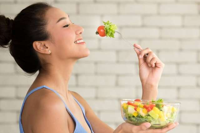Person holding a fork with lettuce and tomato above a glass bowl of mixed vegetable salad.