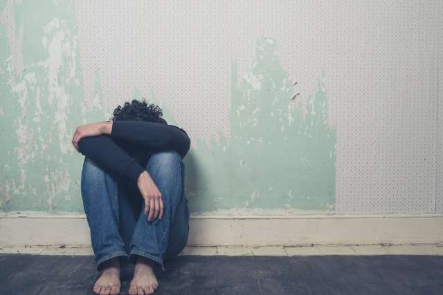 A dejected young man sits on the floor and leans against a peeling wall with his head buried in arms