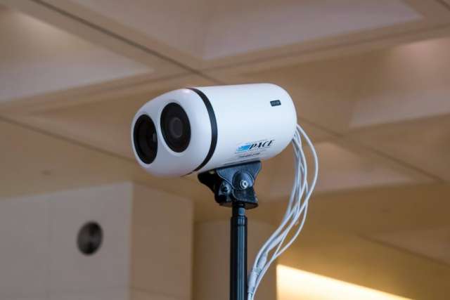 White dual-lens thermal camera on a tripod with cables, used for temperature screening indoors.