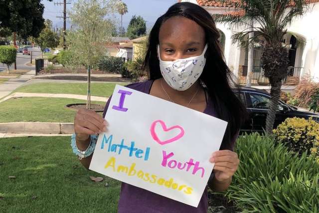 Girl in a mask holds a colorful sign reading "I ❤️ Mattel Youth Ambassadors" while standing outside near grass and palm trees.