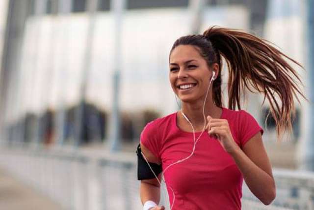 Person jogging outdoors wearing a red shirt, earphones, and an armband, with hair tied back and swinging while running.