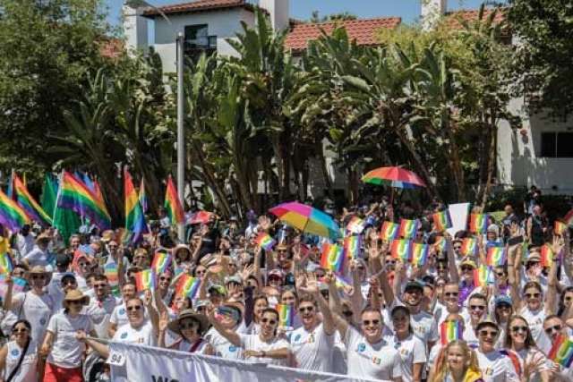 Large crowd at a pride parade holding rainbow flags and umbrellas, with trees and buildings in the background.