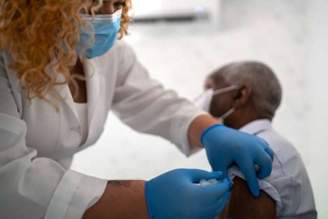 Healthcare worker wearing gloves and a mask administers a vaccine injection to a person’s upper arm in a clinical setting.