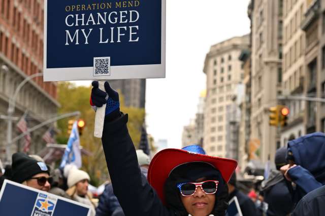 Person wearing a red hat and sunglasses holds a sign reading "Operation Mend CHANGED MY LIFE" at a parade.