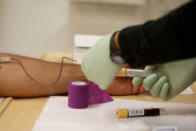 A medical professional's gloved hands draw blood from a patient's arm. A tourniquet and purple bandage are visible near the injection site.