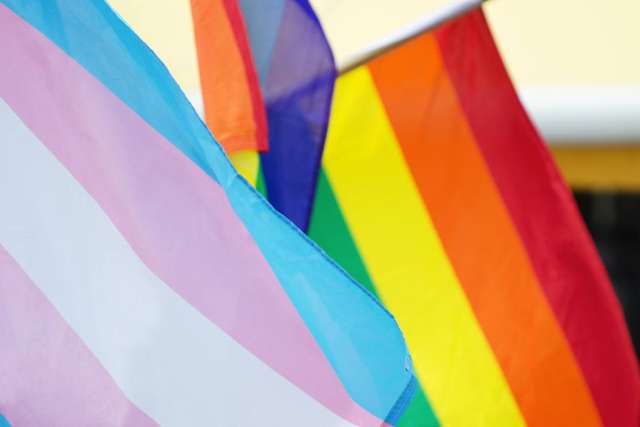 A close-up of the Transgender Pride Flag (blue, pink, white) and the Rainbow Pride Flag (red, orange, yellow, green, blue, purple) flying together.