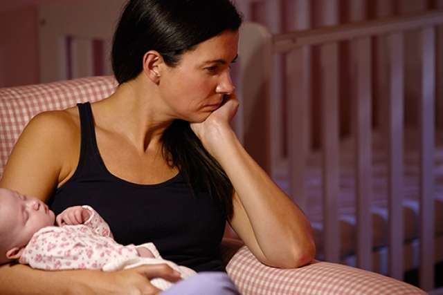 Seated woman holding infant near a crib, looking away — Baby Blues