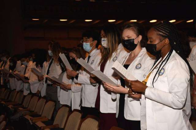 A group of medical students wearing white coats and masks stand and read from papers during a ceremony.