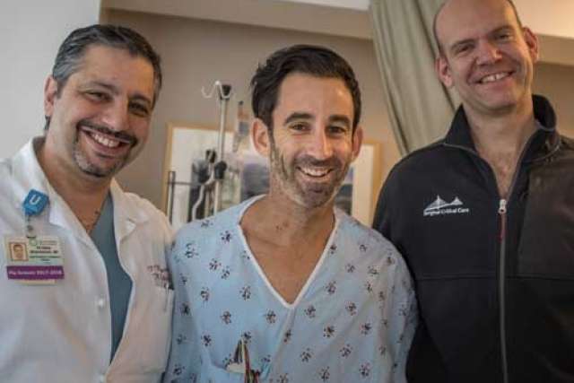 Patient in a hospital gown standing between two people, one in a white medical coat and the other in a dark jacket, in a clinical setting.