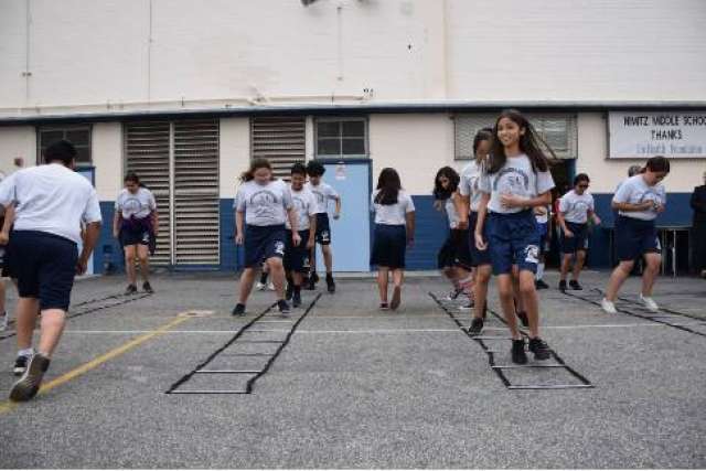 Middle school students in white shirts and blue shorts doing a ladder drill in a school yard.