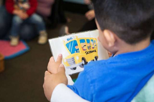 A child in a blue shirt holds a coloring page of a yellow school bus. The child's back is to the camera, with other children blurred in the background.