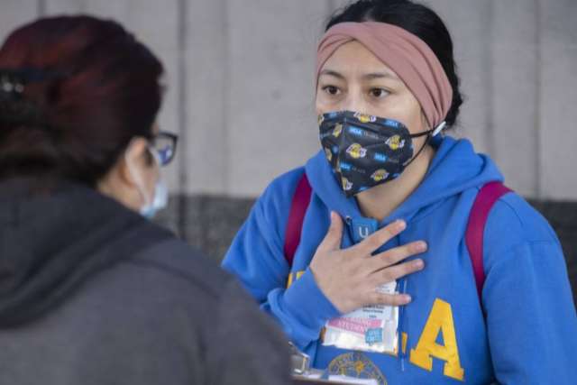 Person wearing a blue UCLA hoodie with a lanyard and backpack, speaking to another individual indoors.