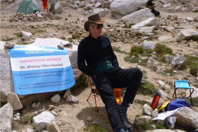 Jonathan Tobis, MD in hiking gear sits beside a sign recruiting Mt. Whitney hikers for a high altitude sickness research study.