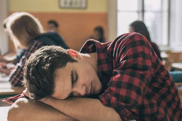 A male student wearing a red and black plaid shirt is asleep with his head resting on his folded arms on a desk in a brightly lit classroom.