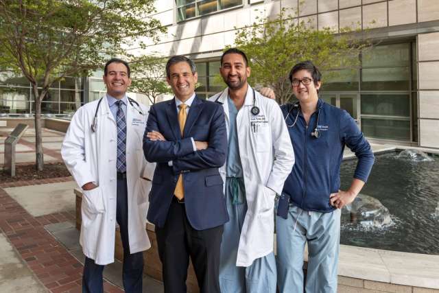 Four men standing side-by-side in front of a modern building. Two men are in white lab coats, one is in a suit, and one is in scrubs.
