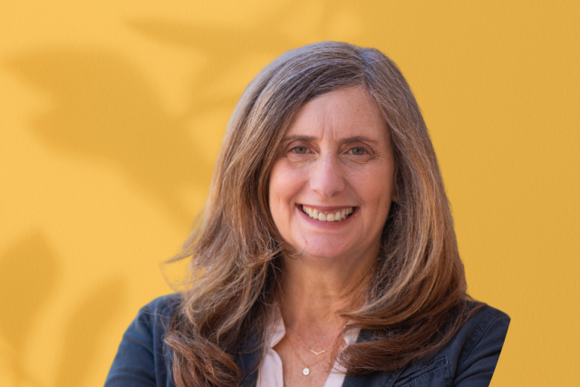 Head-and-shoulders portrait of a woman in a blazer with arms crossed on a yellow background