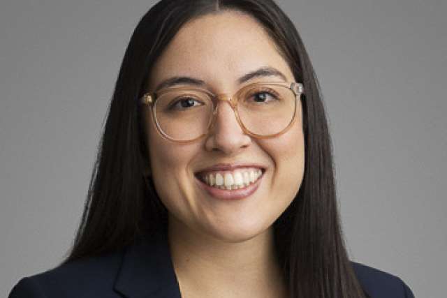 Smiling woman with long dark hair and glasses in a navy blazer on gray background.