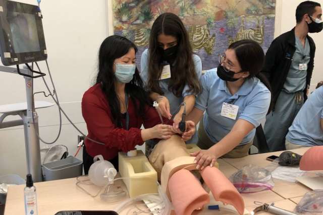 Three allied health students practicing on a medical training mannequin, wearing masks.