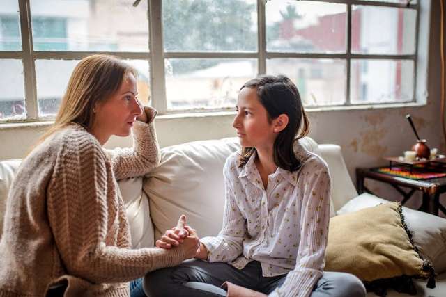 A mother and daughter sit on a white couch by a large window, holding hands and looking at each other seriously during a conversation.